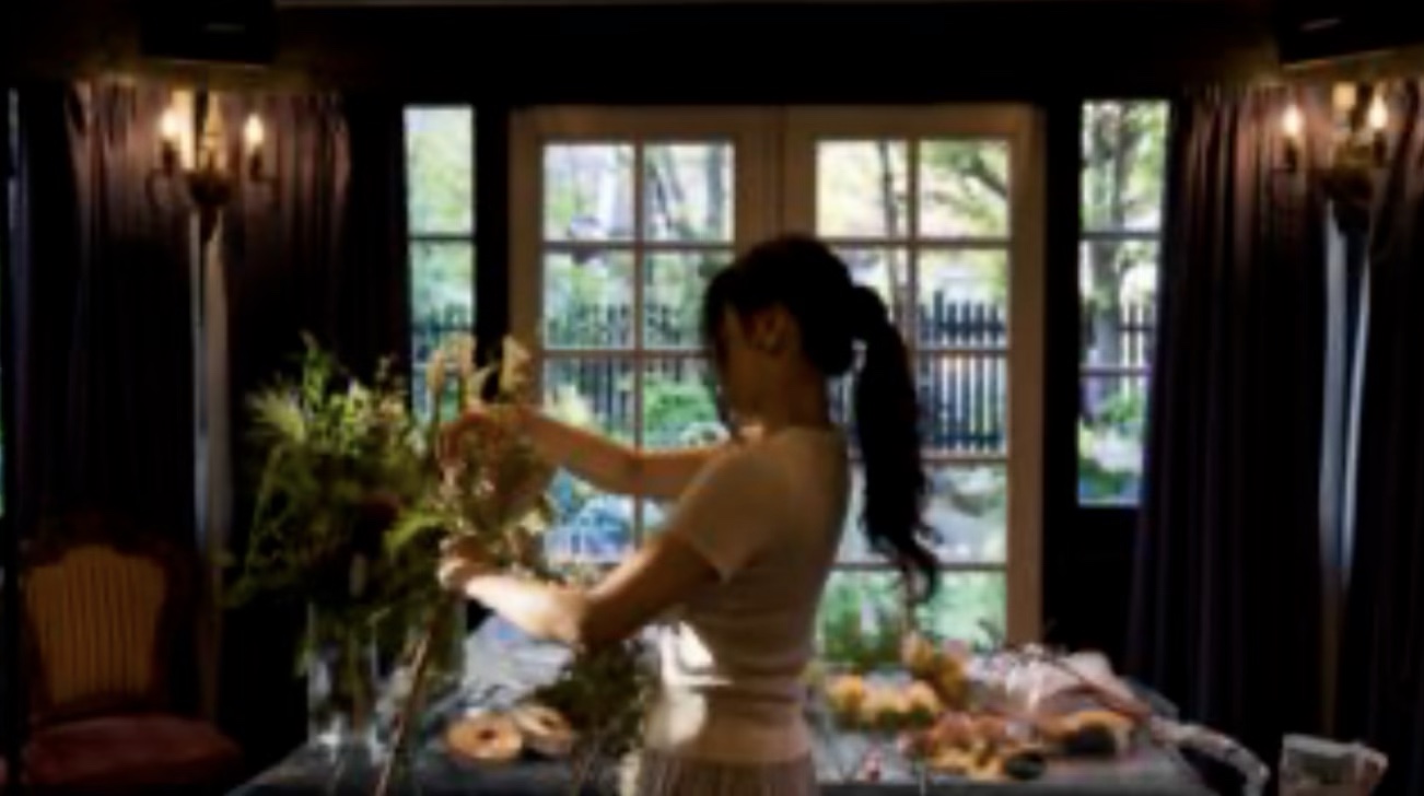 Rinko arranging flowers at a worktable in front of a large window.